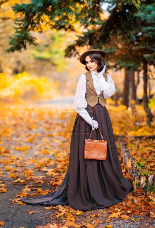 Schöne Frau in Vintage-Kleid, Spitzenbluse und Hut mit Schleier steht in der Nähe von retro braunen Auto auf herbstlichem Hintergrund. Elegante Dame in Handschuhen posiert auf der Natur.Warmes Kunstwerk.