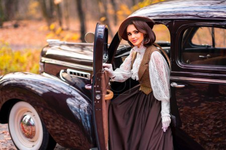 Téléchargez les photos : Belle femelle en robe vintage, chemisier en dentelle et chapeau avec voile assis dans une voiture rétro brune sur fond d'automne. Élégante dame en gants posant sur la nature.Travail d'art chaud. - en image libre de droit