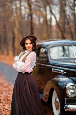 Photo for Beautiful female in vintage  dress, lace blouse and hat with veil standing near retro brown car on autumn background. Elegant lady in gloves posing on nature.Warm art work. - Royalty Free Image