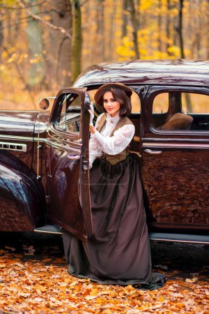 Schöne Frau in Vintage-Kleid, Spitzenbluse und Hut mit Schleier sitzt im retro braunen Auto auf herbstlichem Hintergrund. Elegante Dame in Handschuhen posiert auf der Natur.Warmes Kunstwerk.