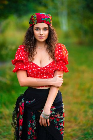 Photo for Pretty brunette curly girl in red gipsy costume and accessories posing on nature with green background and bonfire. Warm art work with queen. - Royalty Free Image