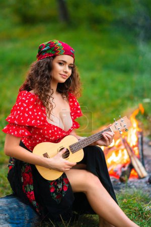 Foto de Linda chica rizada morena en traje de gitano rojo y accesorios posando sobre fondo verde naturaleza. Reina tocando en la guitarra. - Imagen libre de derechos