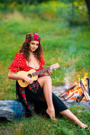 Foto de Linda chica rizada morena en traje de gitano rojo y accesorios posando sobre fondo verde naturaleza. Reina tocando en la guitarra. - Imagen libre de derechos