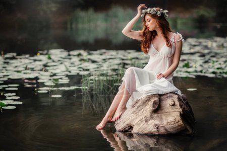 Schöne rothaarige Mädchen in weißen Vintage-Kleid und Blumenkranz sitzt am Ufer des Flusses. Märchenhafte Geschichte. Warmes Kunstwerk.
