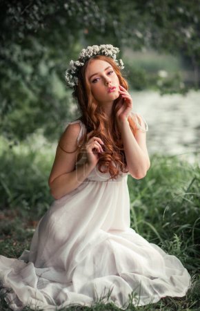 Beautiful red haired girl in white vintage dress and wreath of flowers standing under the tree and looking at camera. Fairytale story. Warm art work.