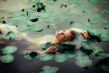 Foto de Beautiful red haired girl in white dress posing in river with water lilies. Fairytale story about  ophelia .Warm art work - Imagen libre de derechos