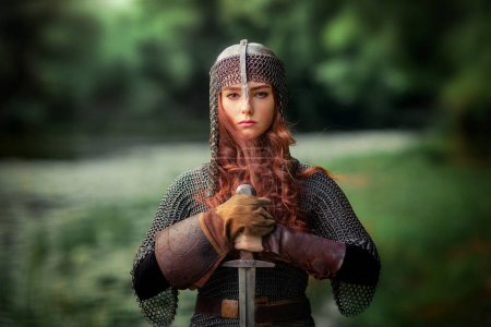 Hermosa chica pelirroja en vestido de armadura medieval de metal con espada de pie en pose guerrera. Historia de cuento de hadas sobre guerrero. Trabajo de arte cálido.