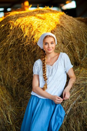 Téléchargez les photos : Belle fille blonde aux cheveux tressés en vêtements ruraux blancs assis sur une paille. Vie à la ferme.Jeune mannequin posant sur un ranch.. - en image libre de droit