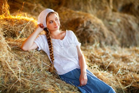 Téléchargez les photos : Belle fille blonde aux cheveux tressés en vêtements ruraux blancs assis sur une paille. Vie à la ferme.Jeune mannequin posant sur un ranch.. - en image libre de droit