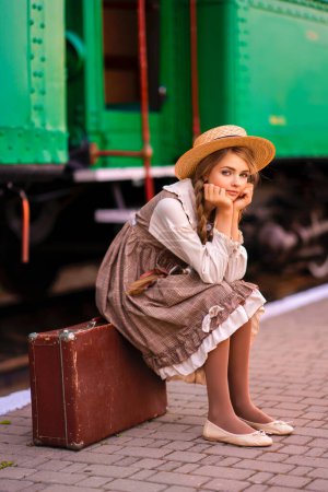 Foto de Retrato de la joven chica trenzada rubia bonita en vestido vintage y sombrero de paja de pie en la plataforma de la estación cerca del tren verde. Sonriente elegancia linda dama con ojos azules posando. Obra de arte sobre viajero. - Imagen libre de derechos