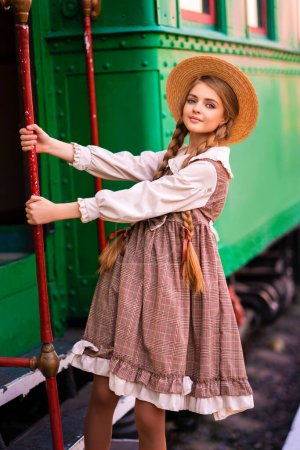 Téléchargez les photos : Portrait de jeune jolie fille blonde tressée en robe vintage et chapeau de paille debout sur la plate-forme de la gare près du train vert. Élégance souriante dame mignonne avec les yeux bleus posant. Oeuvre d'art sur voyageur. - en image libre de droit