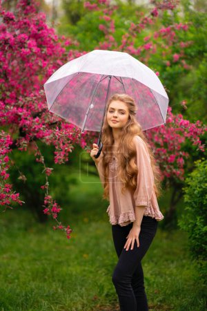 Photo for Pretty young blonde girl with long curly hair in vintage lace blouse and  green cloak standing in spring park near pink blossom flowers. Tenderness romantic  model posing with transparent umbrella. - Royalty Free Image