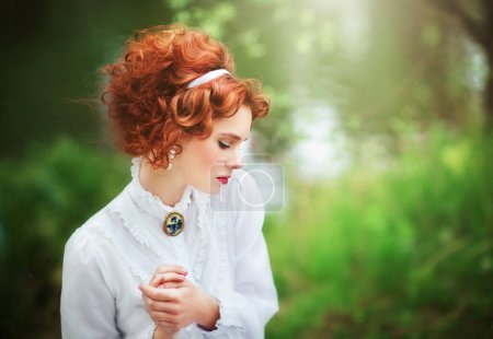 Téléchargez les photos : Belle fille aux cheveux rouges en robe longue vintage blanche assise sur une herbe près de la rivière.Oeuvre d'art de femme romantique avec un livre et un parapluie en dentelle. - en image libre de droit