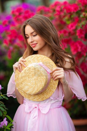 Belle fille en robe vintage rose et chapeau de paille debout près de fleurs colorées. Oeuvre d'art de femme romantique. Jolie modèle de tendresse posant et regardant la caméra.