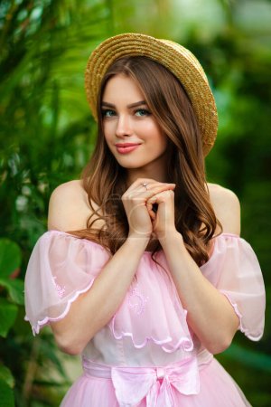 Téléchargez les photos : Belle fille en robe vintage rose et chapeau de paille debout sur fond vert avec des plantes. Oeuvre d'art de femme romantique. Jolie modèle de tendresse posant et regardant la caméra. - en image libre de droit