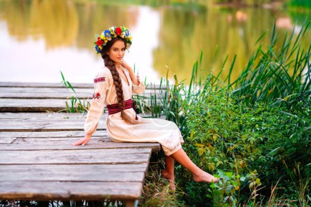 Hermosa chica de pelo largo trenzado en vestido tradicional ucraniano y corona posando en la naturaleza verde cerca del río. Retrato de mujer elegante atractiva joven sobre fondo cálido colorido.
