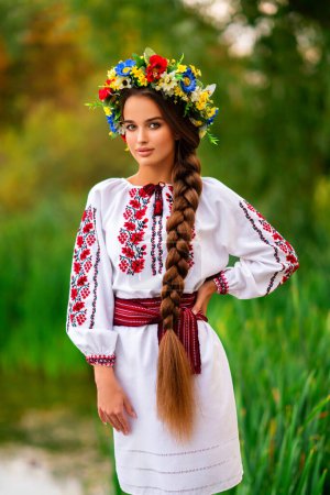 Téléchargez les photos : Belle fille cheveux longs tressés en robe traditionnelle ukrainienne et couronne posant sur la nature verte près de la rivière. Portrait de jeune femme élégante attrayante sur fond chaud coloré. - en image libre de droit