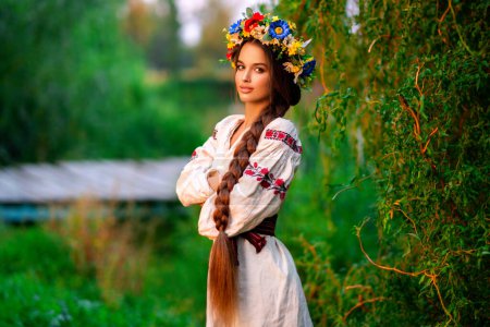 Hermosa chica de pelo largo trenzado en vestido tradicional ucraniano y corona posando en la naturaleza verde cerca del río. Retrato de mujer elegante atractiva joven sobre fondo cálido colorido.