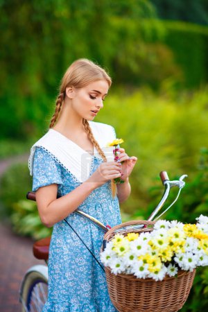 Photo for Beautiful young blonde smiling girl with short blue dress standing near white bicycle with basket of huge bouquet of chamomiles.Summer lady enjoying the nature outdoor.Green background with flowers. - Royalty Free Image