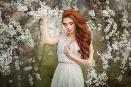 Photo for Spring Beautiful romantic red haired girl in white  lace dress standing in blooming garden. Dreaming young model looking down. - Royalty Free Image