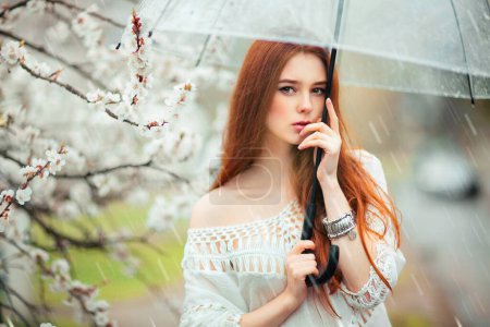 Foto de Primavera Hermosa chica romántica de pelo rojo en vestido de encaje blanco y jeans de pie cerca del árbol floreciente con umbrella.Young modelo transparente cerca de la carretera bajo la lluvia mirando a la cámara
. - Imagen libre de derechos