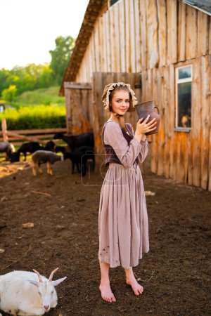 Beautiful young rural girl in medieval dress and bonnet hat posing on a ranch with jug of milk. Pretty blonde model in countryside art photo. 