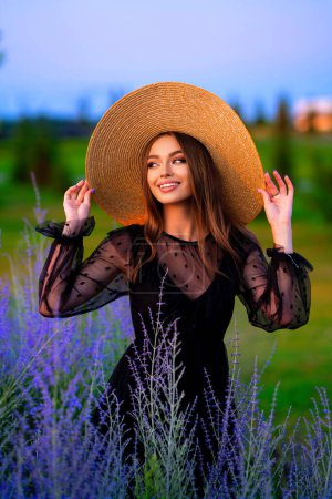 Foto de Retrato de una hermosa chica en un vestido negro transparente y un gran sombrero de paja de pie en el campo de color lavanda. Obra de arte de mujer sonriente romántica . - Imagen libre de derechos