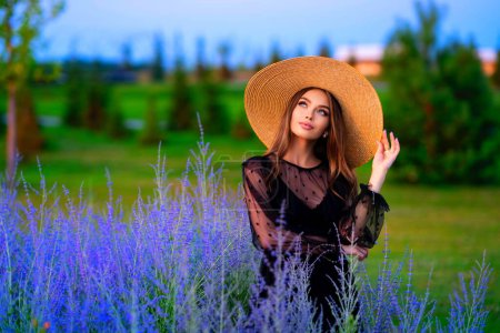 Photo for Portrait of a Beautiful girl in a black transparent dress and straw big hat standing in colorful lavender field. Art work of romantic woman . - Royalty Free Image