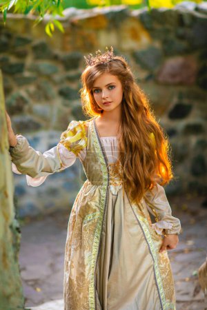 Foto de Hermosa princesa rubia joven en vestido medieval de oro y pequeña corona. Linda reina posando cerca de piedra castle.Warm obra de arte. - Imagen libre de derechos