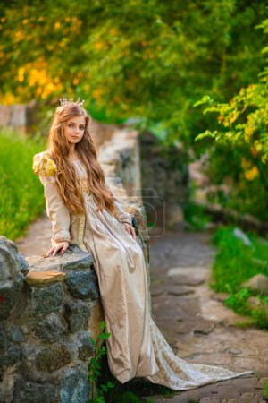 Foto de Hermosa princesa rubia joven en vestido medieval de oro y pequeña corona. Linda reina posando cerca de piedra castle.Warm obra de arte. - Imagen libre de derechos