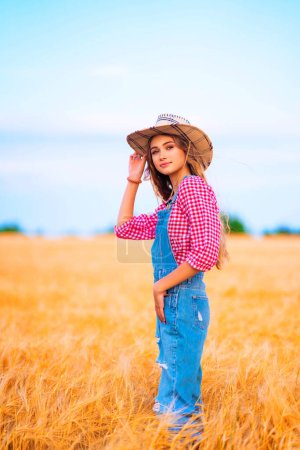 Téléchargez les photos : Jeune jolie fille blonde posant dans le domaine du blé.Belle cowgirl élégante en chapeau de paille et salopettes en denim sur des photos de campagne colorées chaudes. - en image libre de droit