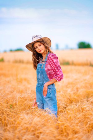 Photo for Young pretty blonde girl posing in field of wheat.Beautiful stylish cowgirl in straw hat and denim overalls  on warm colorful countryside pictures. - Royalty Free Image