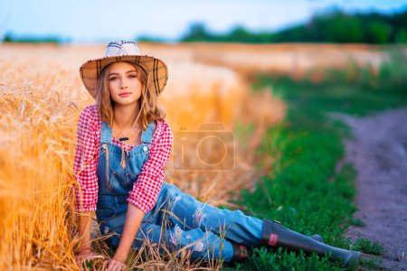 Foto de Joven chica rubia bonita posando en el campo de wheat.Beautiful vaquera con estilo en sombrero de paja y overoles de mezclilla en cálidas imágenes coloridas del campo. - Imagen libre de derechos