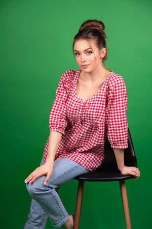 Téléchargez les photos : Jolie jeune fille en chemise à carreaux et jeans posant sur chaise sur fond vert isolé.Studio travailler avec un modèle attrayant. - en image libre de droit