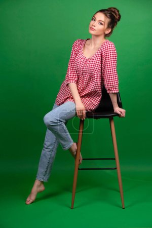 Foto de Chica bastante joven en camisa a cuadros y jeans posando en silla sobre fondo verde aislado. trabajo de estudio con modelo atractivo. - Imagen libre de derechos