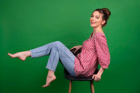 Téléchargez les photos : Jolie jeune fille en chemise à carreaux et jeans posant sur chaise sur fond vert isolé.Studio travailler avec un modèle attrayant. - en image libre de droit