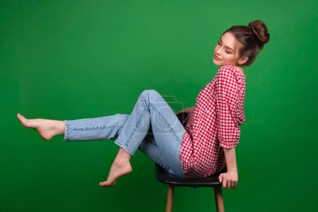 Foto de Chica bastante joven en camisa a cuadros y jeans posando en silla sobre fondo verde aislado. trabajo de estudio con modelo atractivo. - Imagen libre de derechos