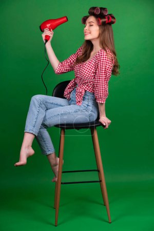 Photo for Pretty young smiling girl in big red curlers drying hair with red dryer. Studio work with attractive model in checkered red shirt and jeans posing  on green background isolated. - Royalty Free Image