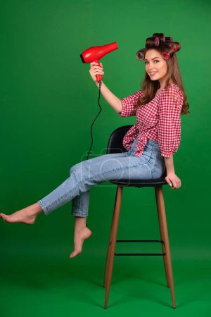 Photo for Pretty young smiling girl in big red curlers drying hair with red dryer. Studio work with attractive model in checkered red shirt and jeans posing  on green background isolated. - Royalty Free Image