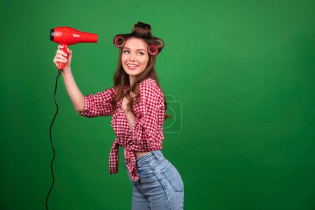 Téléchargez les photos : Jolie jeune fille souriante en gros bigoudis rouges séchant les cheveux avec sèche-cheveux rouges. Travail en studio avec un modèle attrayant en chemise rouge à carreaux et jeans posant sur fond vert isolé. - en image libre de droit