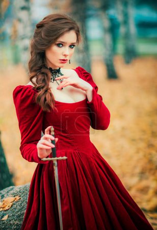 Portrait of magnificent Fashion gothic girl standing in autumn forest .Fantasy art work.Amazing red haired model in claret dress with a sword .Fairytale about young princess-warrior. 