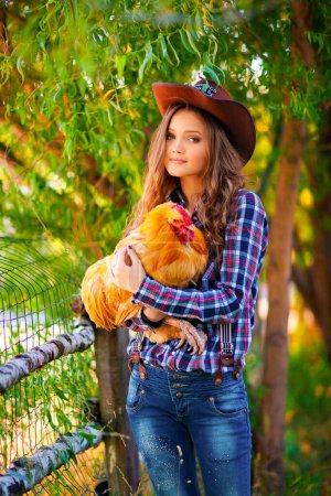 Téléchargez les photos : Jeune belle fille dans un chapeau de cow-boy, jeans et chemise à carreaux posant avec la poule. Jolie modèle de cheveux longs debout près de foin loft outdoor .Country style.Art travail. - en image libre de droit