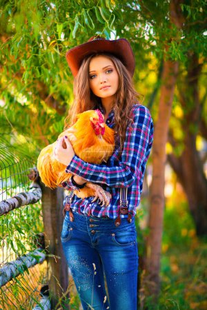 Photo for Young beautiful girl in a cowboy hat, jeans and checkered shirt posing with hen.  Pretty long hair model standing near hayloft outdoor .Country style.Art work. - Royalty Free Image