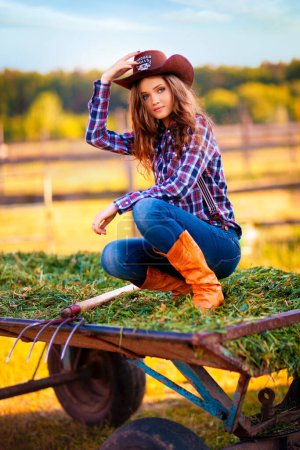 Photo for Young beautiful girl in a cowboy hat, jeans and checkered shirt posing and looking at camera.  Pretty long hair model standing near hayloft outdoor .Country style.Art work. - Royalty Free Image