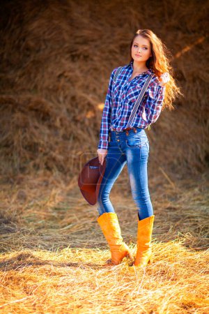 Photo for Young beautiful girl in a cowboy hat, jeans and checkered shirt posing and looking at camera.  Pretty long hair model standing near hayloft outdoor .Country style.Art work. - Royalty Free Image