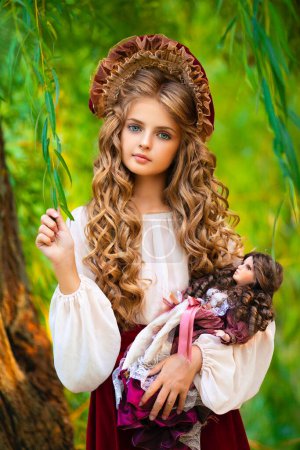 Photo for Beautiful young girl in vintage dress and cute red hood outdoor. Art work of romantic girl with a doll. Pretty tenderness blonde model with huge blue eyes. - Royalty Free Image