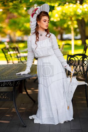 Beautiful red haired girl in white long vintage dress and hat sitting near vintage table on sunny background.Pretty tenderness model with perfect hairstyle and make up posing with umbrella. Art work and fairytale