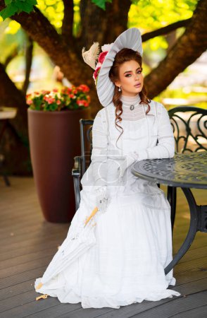 Schöne rothaarige Mädchen in weißen langen Vintage-Kleid und Hut sitzt in der Nähe Vintage-Tisch auf sonnigem Background.Pretty Zartheit Modell mit perfekter Frisur und Make-up posiert mit Regenschirm. Kunstwerk und Märchen