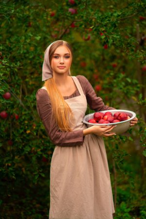 Téléchargez les photos : Portrait de belle fille blonde en robe médiévale et tablier aux pommes rouges. Jeune travailleur debout dans le jardin avec bol de pommes. Travail d'art coloré chaud. - en image libre de droit
