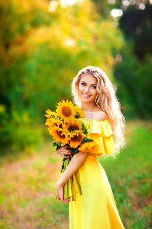 Photo for Beautiful young blonde girl in yellow dress enjoying nature. Happy Smiling female standing in green forest with sunflowers. Warm photo. Art work. - Royalty Free Image